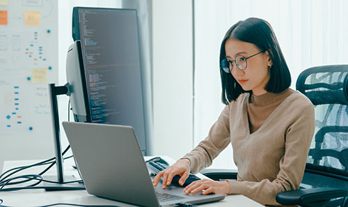 a woman sitting at a desk typing on a laptop keyboard, a large screen in front of her
