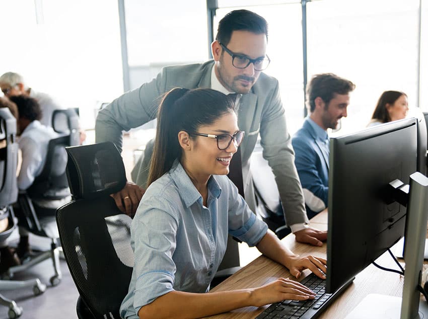 A professional setting where a woman is working on a computer while a man stands behind her, providing support.