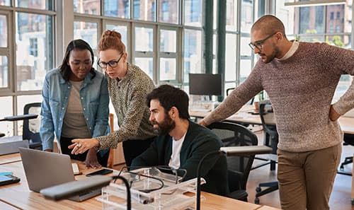 Four people, around a table in a light filled office. One is pointing at something on a laptop screen.