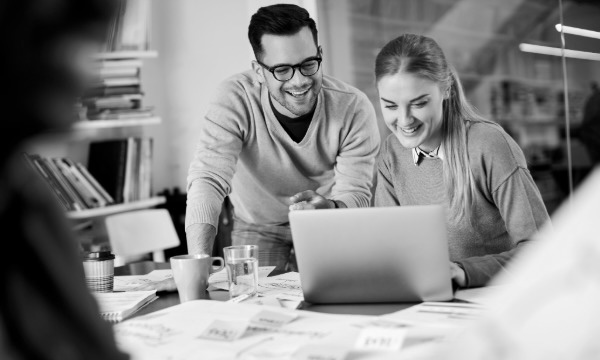 Two coworkers smiling while reviewing something on a laptop at a desk covered with papers in an office.