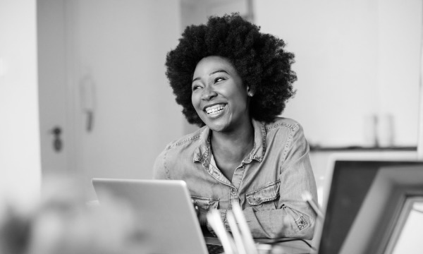 Smiling woman with natural afro sitting at a desk with a laptop in a bright office.